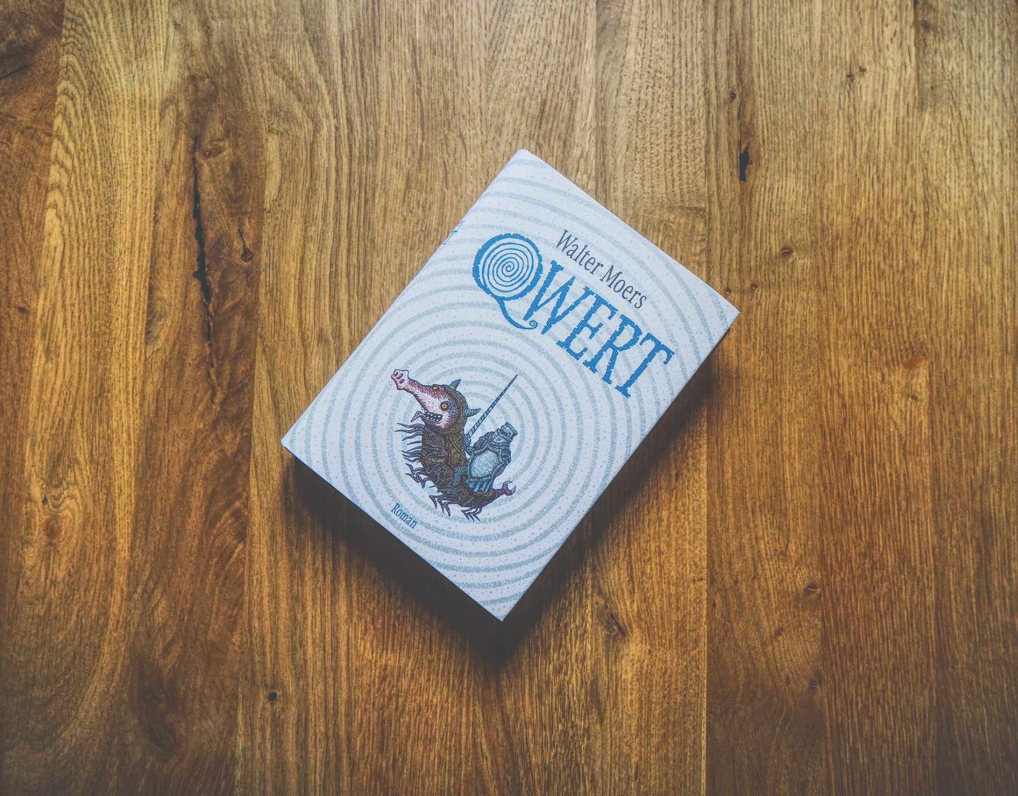 Auf einer hölzernen Unterlage liegt ein Buch mit dem Titel "Qwert" von Walter Moers, das auf dem Einband ein illustriertes Wesen mit Horn und Schwanz zeigt.