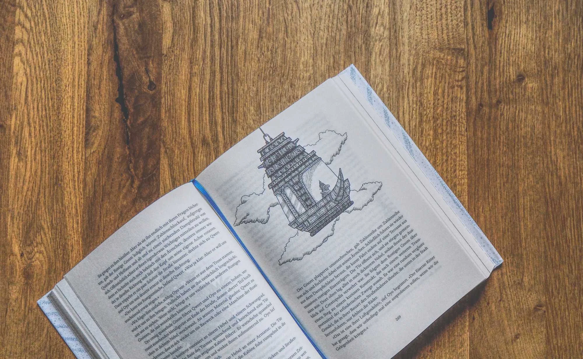 Ein aufgeschlagenes Buch liegt auf einer hölzernen Unterlage und zeigt eine Schwarz-Weiß-Illustration eines fliegenden Schiffes über dem Text.