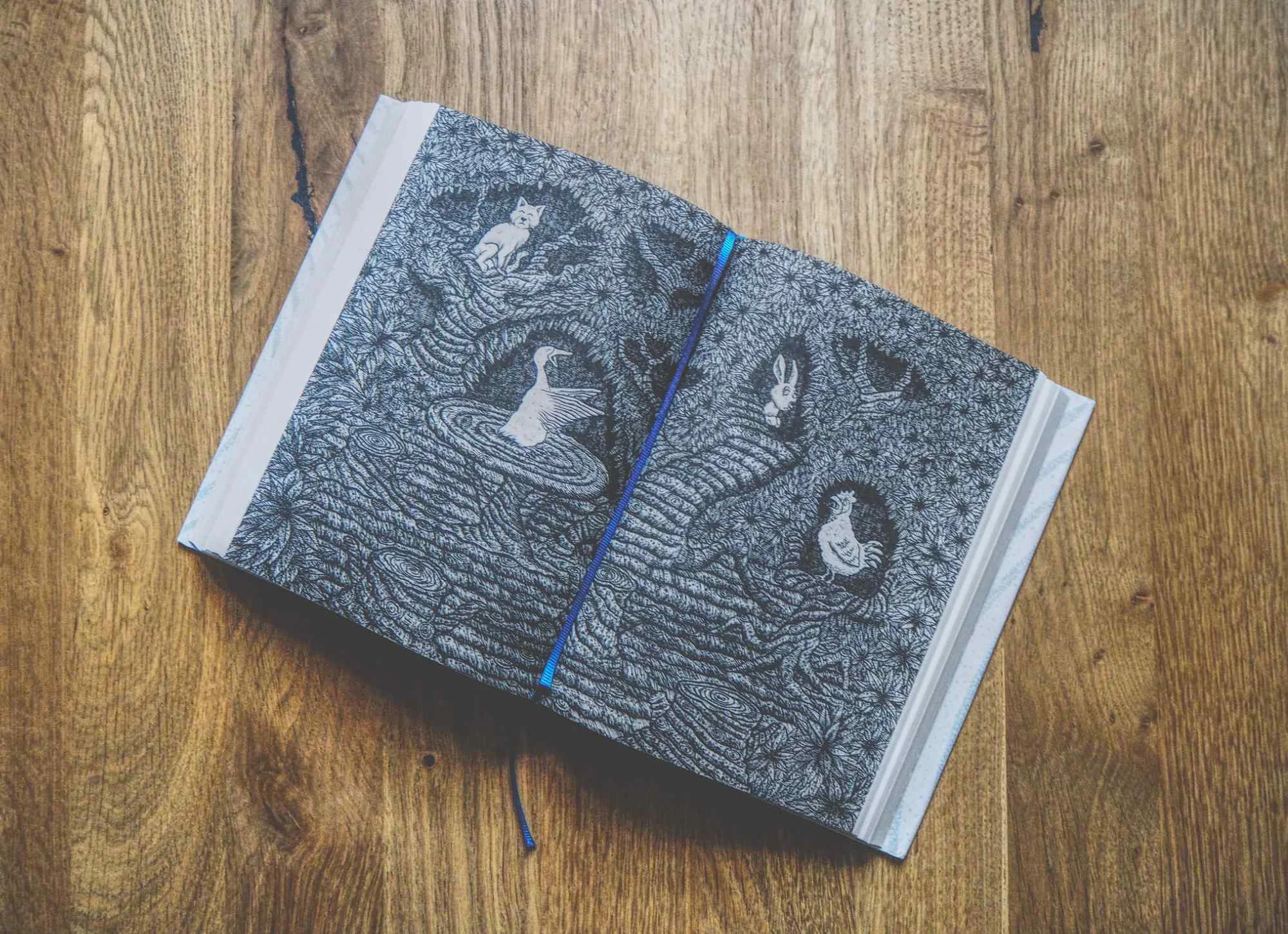 Ein aufgeschlagenes Buch mit aufwändigen Schwarz-Weiß-Illustrationen von Tieren und Figuren in einer Waldumgebung, die auf einer Holzfläche platziert sind.