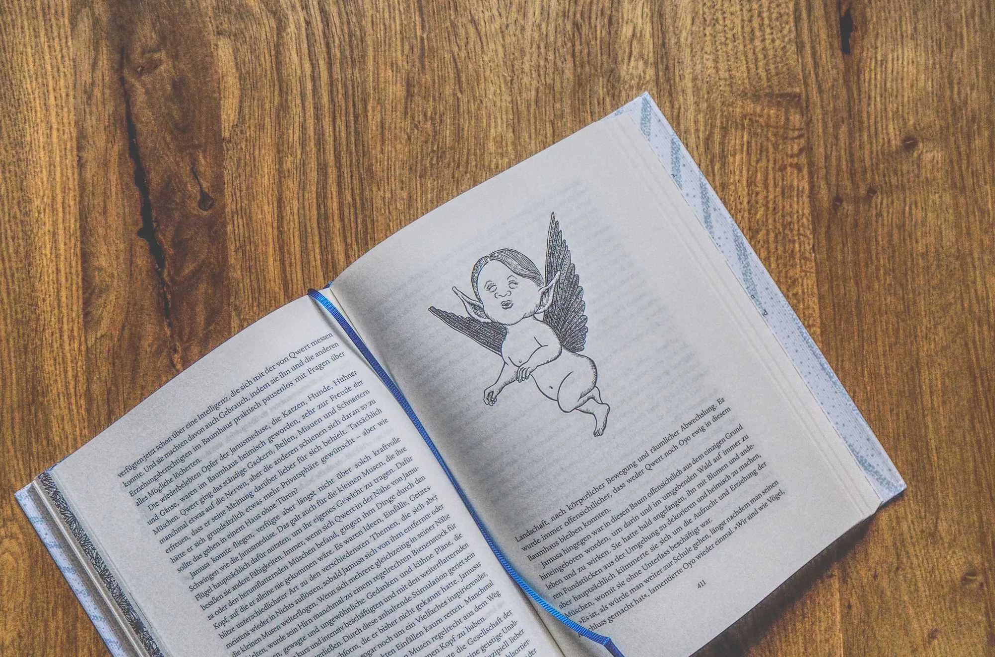 Ein aufgeschlagenes Buch auf einer hölzernen Oberfläche zeigt eine Seite mit Text und die Illustration einer geflügelten Engelsfigur.