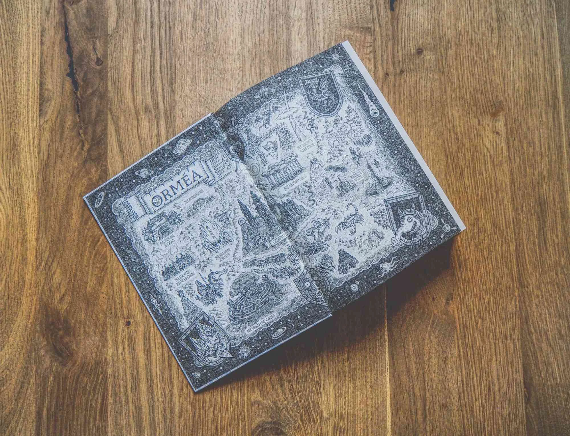 Ein aufgeschlagenes Buch mit einer detailliert illustrierten Fantasiekarte mit der Aufschrift "ORMEA", die auf einer hölzernen Unterlage liegt.