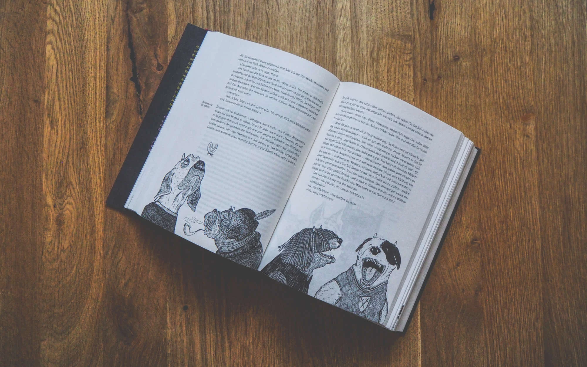 Auf einem Holztisch liegt ein aufgeschlagenes Buch mit Text auf beiden Seiten und Schwarz-Weiß-Illustrationen von bekleideten Tieren am unteren Rand.