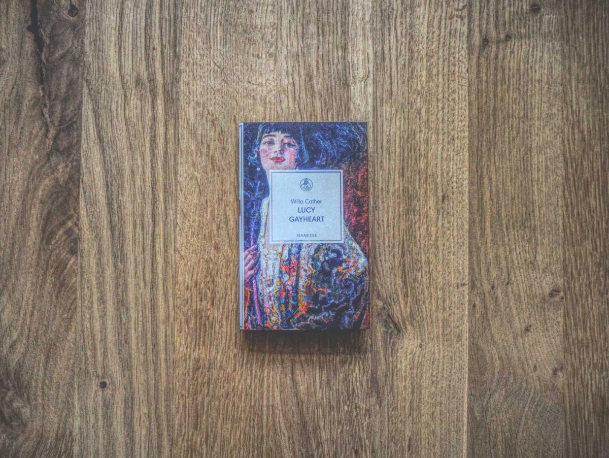 Ein Buch mit dem Titel „Lucy Gayheart“ von Willa Cather mit einem illustrierten Cover, das eine Frau zeigt, die auf einer Holzoberfläche platziert ist.