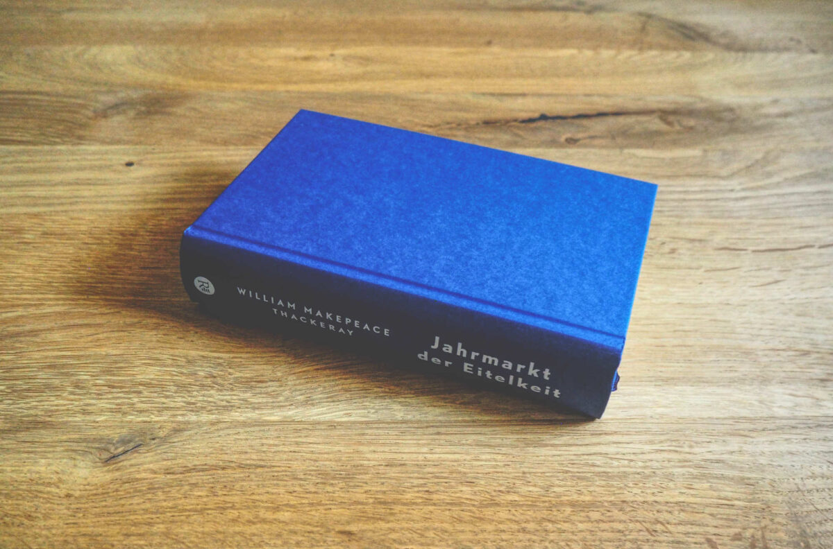 Ein blaues, gebundenes Buch mit dem Titel „Jahrmarkt der Eitelkeit“ von William Makepeace Thackeray liegt geschlossen auf einer Holzfläche.