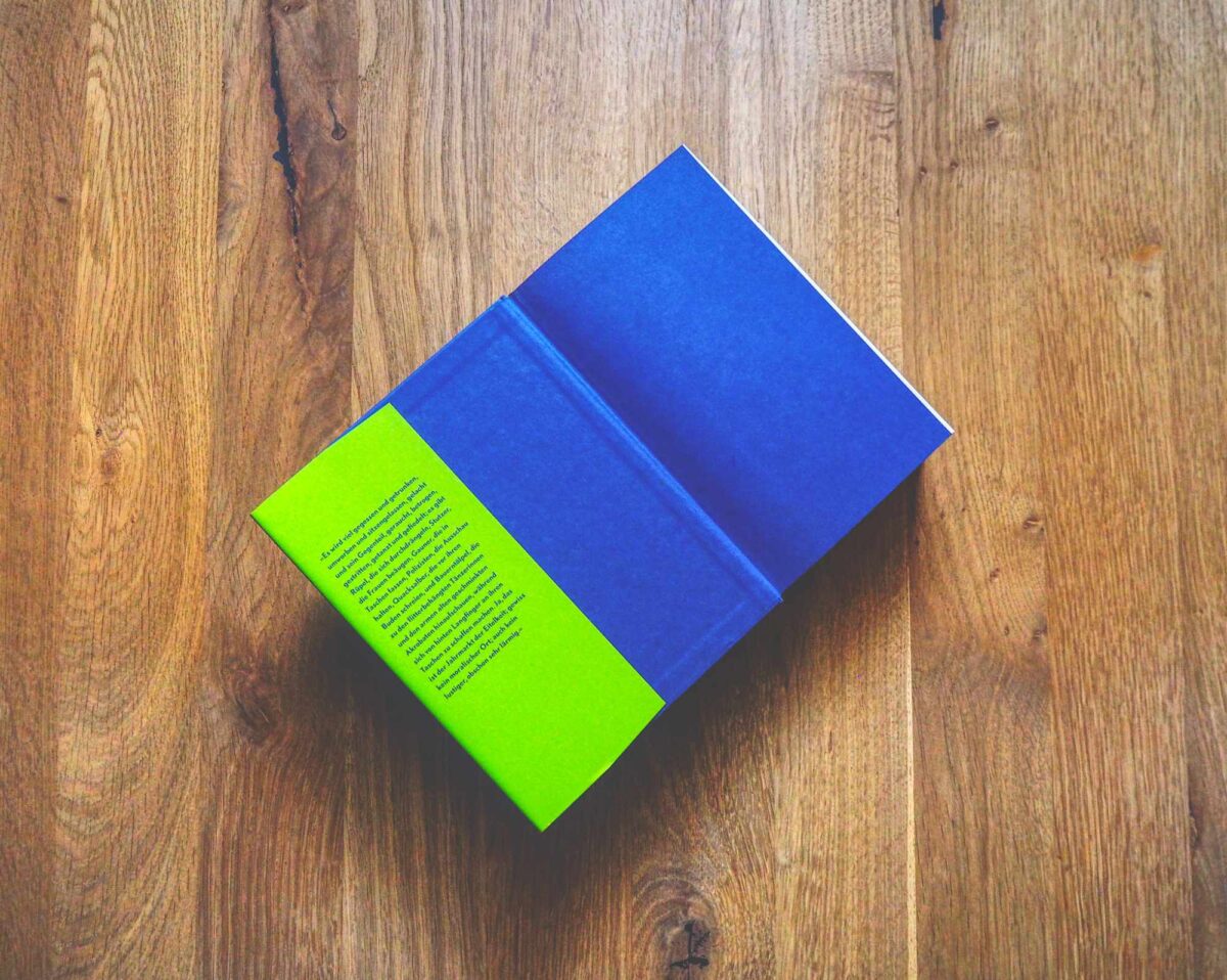 Ein geschlossenes Buch mit einem blau-grünen Einband liegt auf einer Holzfläche, von oben betrachtet.