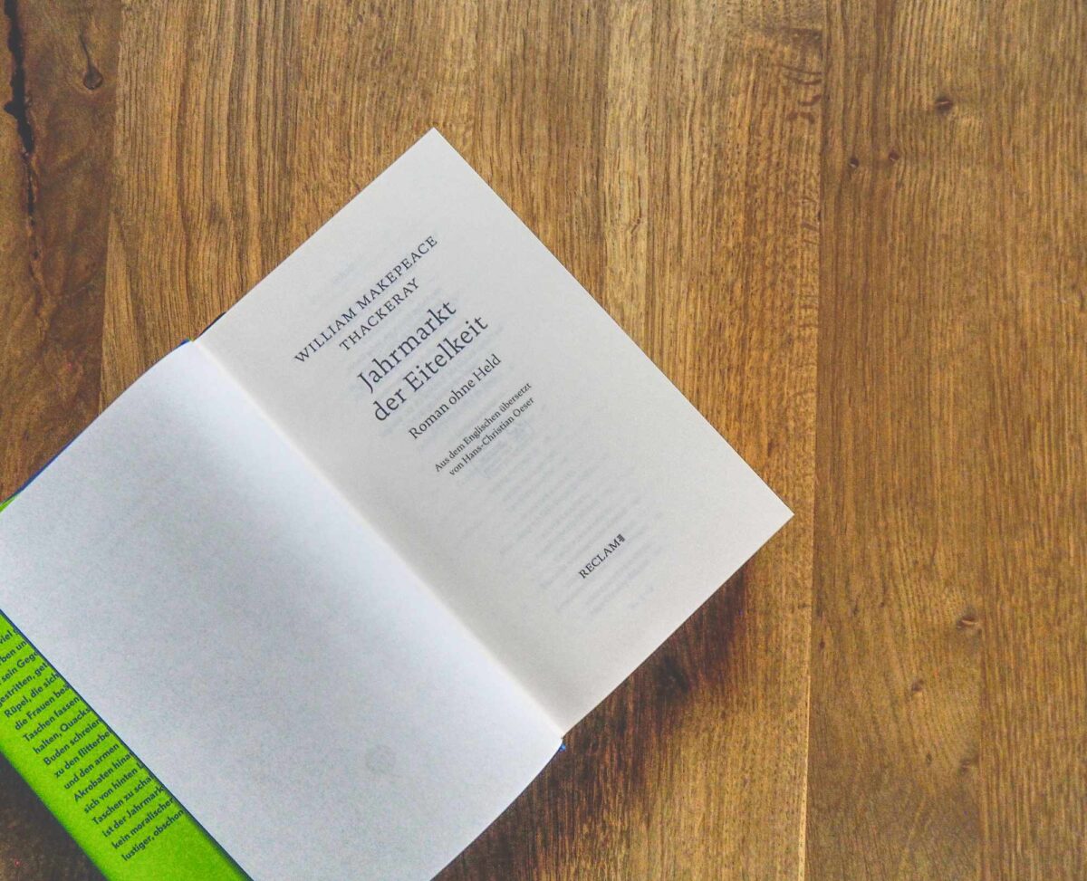 Ein aufgeschlagenes Buch mit dem Titel „Jahrmarkt der Eitelkeit“ von William Makepeace Thackeray liegt auf einer Holzfläche, wobei die Titelseite in deutscher Sprache zu sehen ist.