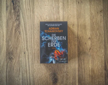Auf einer Holzfläche liegt ein Buch mit dem Titel „Die Scherben der Erde“ von Adrian Tchaikovsky. Der Einband ist mit kosmischen Motiven in Blau und Rot gestaltet.