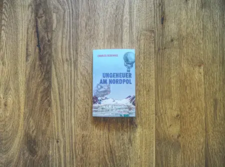Buch mit dem Titel "Ungeheuer am Nordpol" von Charles Derrennes auf einer Holzoberfläche. Auf dem Einband sind Berge, ein Heißluftballon und eisige Landschaften abgebildet.