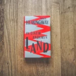 Auf einer Holzfläche liegt eine deutsche Ausgabe von Ernest Hemingways Buch „In einem anderen Land“.