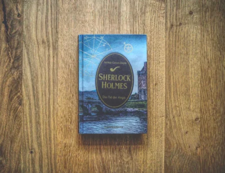 Ein gebundenes Buch mit dem Titel „Sherlock Holmes: Das Tal der Angst“ von Arthur Conan Doyle liegt auf einer Holzfläche. Auf dem Cover ist ein altes Gebäude neben einem Fluss abgebildet.