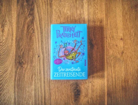Auf einer Holzfläche liegt ein Buch mit dem Titel „Der zertstreute Zeitreisende“ von Terry Pratchett mit illustriertem Cover.