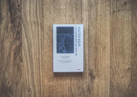 Ein Buch mit dem Titel „Der Wanderer auf dem Eis“ von Volter Kilpi liegt auf einer Holzoberfläche. Auf dem Cover ist ein verschneiter Pfad abgebildet.